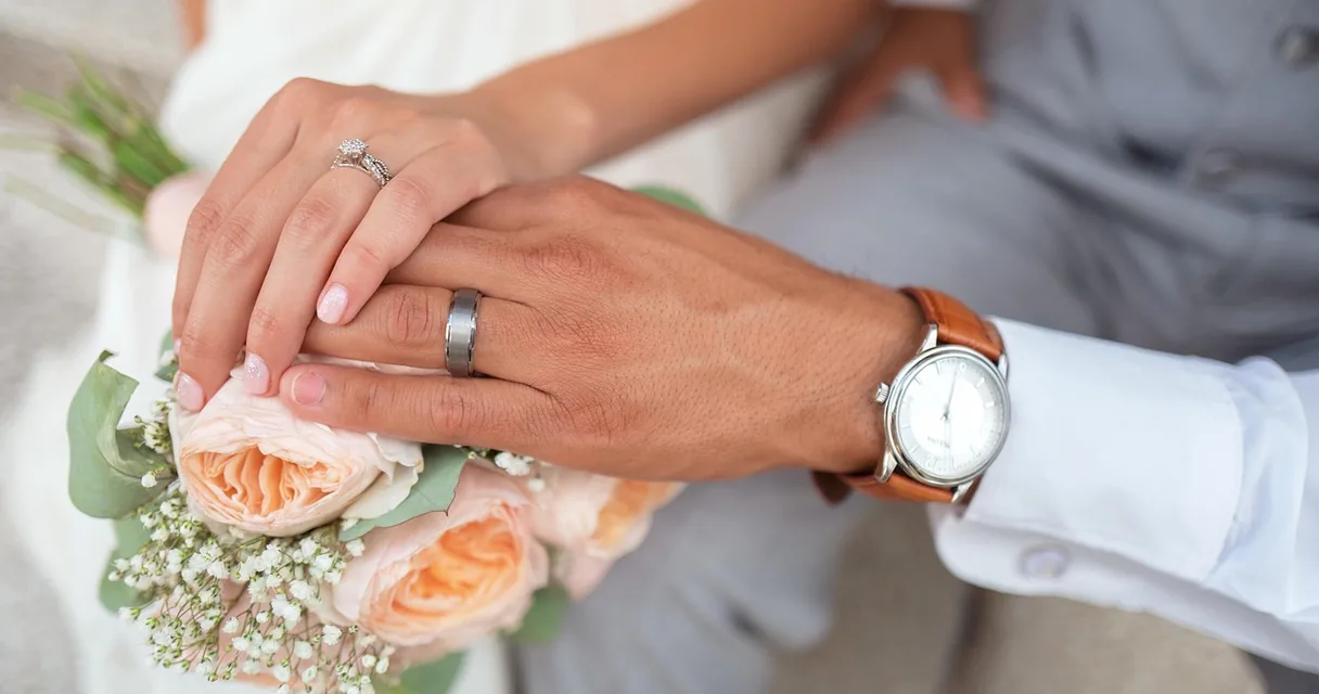 Mãos de um casal com alianças sobre um buquê de rosas, ilustrando sonhar com casamento